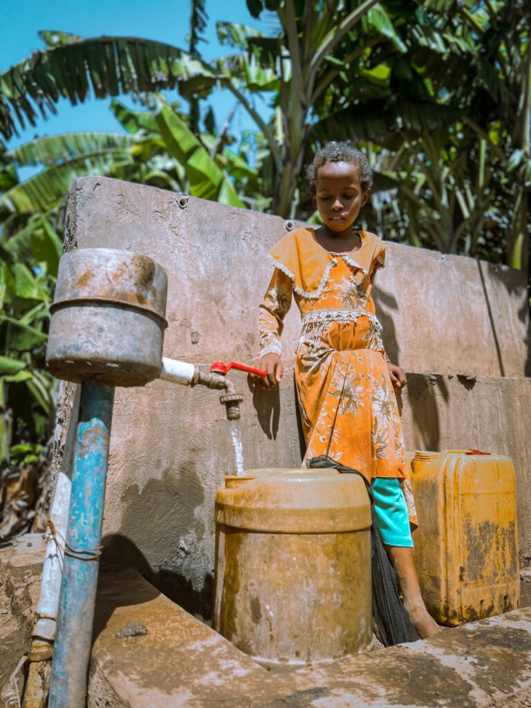 pexels-photo-13799233-13799233 A young girl in vibrant clothing draws water from a well in Afgoye, Somalia, surrounded by lush vegetation.